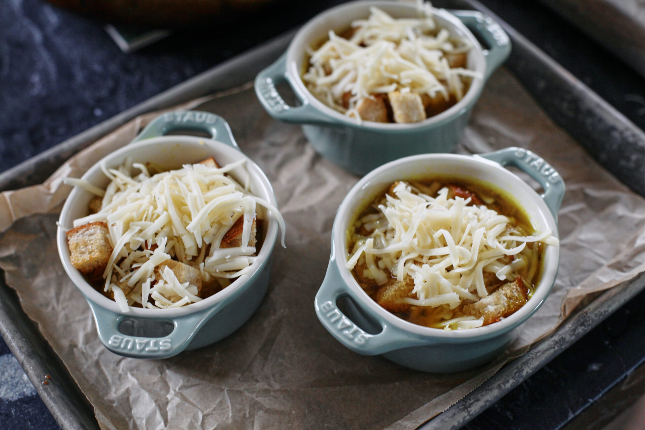 oven-safe crocks arranged on a sheet pan. They're filled with soup and topped with croutons and grated cheese