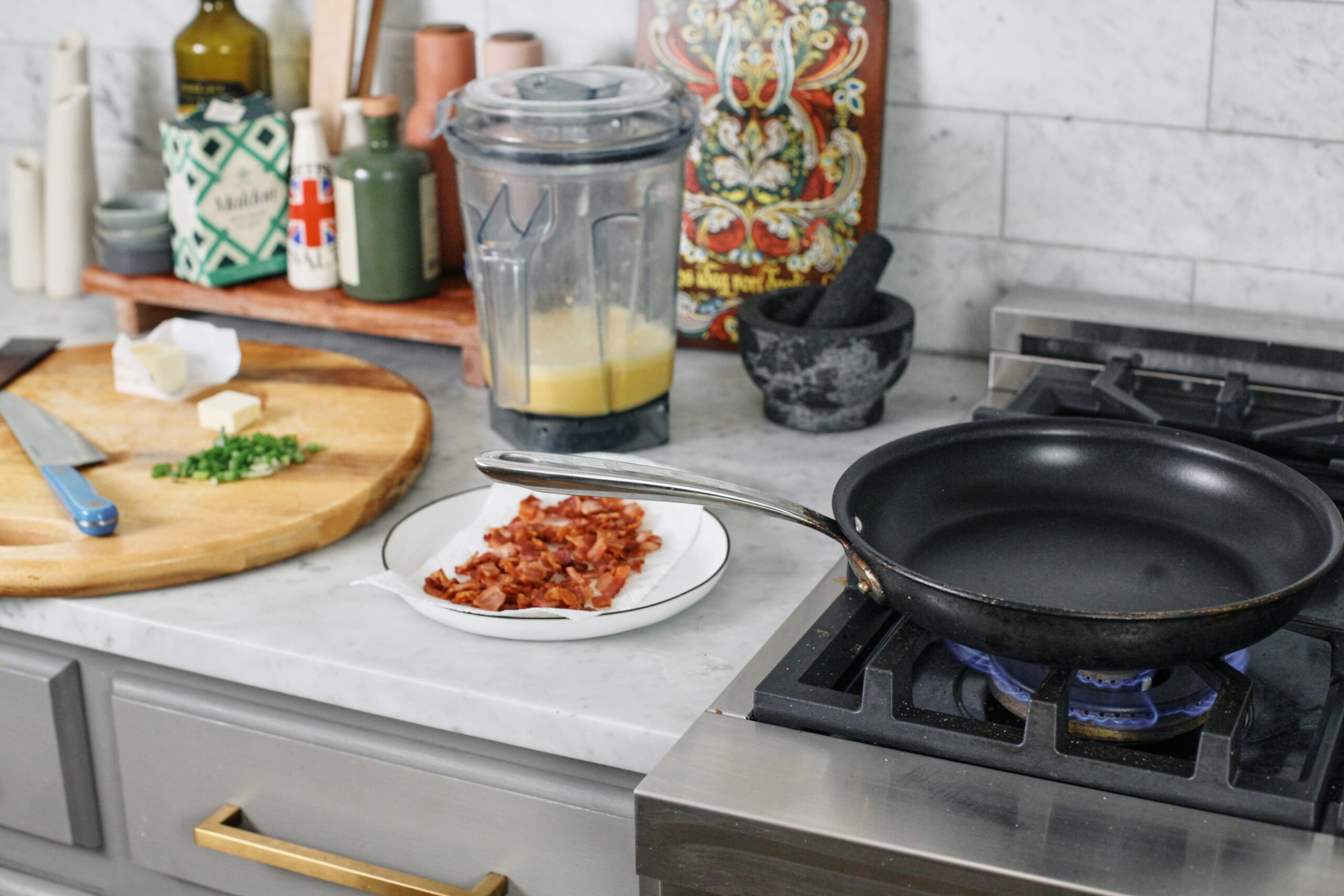 Kitchen counter and stovetop. There's a cutting board, blender, and plate of cooked bacon on the countertop, and an empty skillet on the stove.