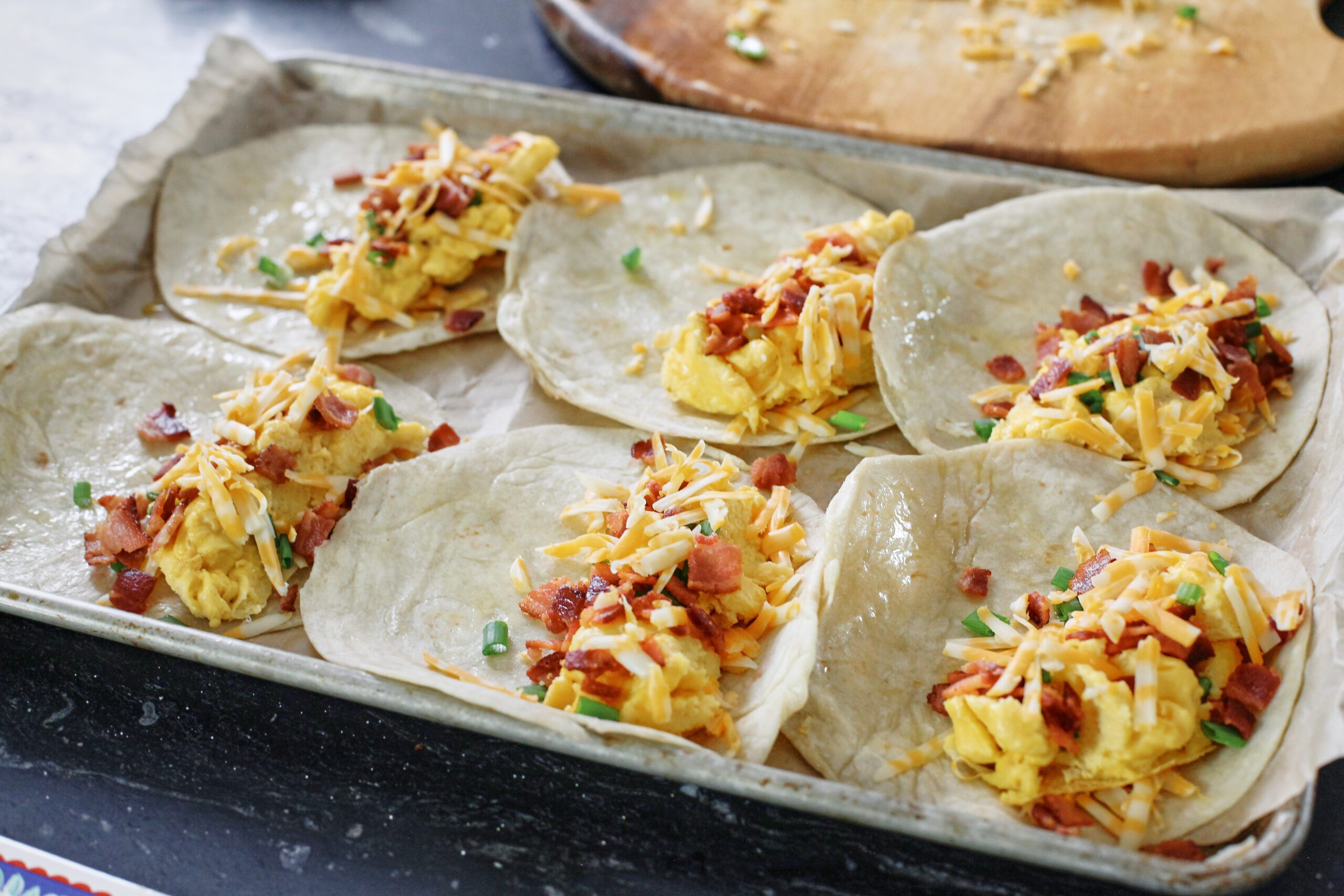 Bacon, eggs, green onion, colby jack shredded cheese on one half of 6 tortillas, arranged on a sheet pan.