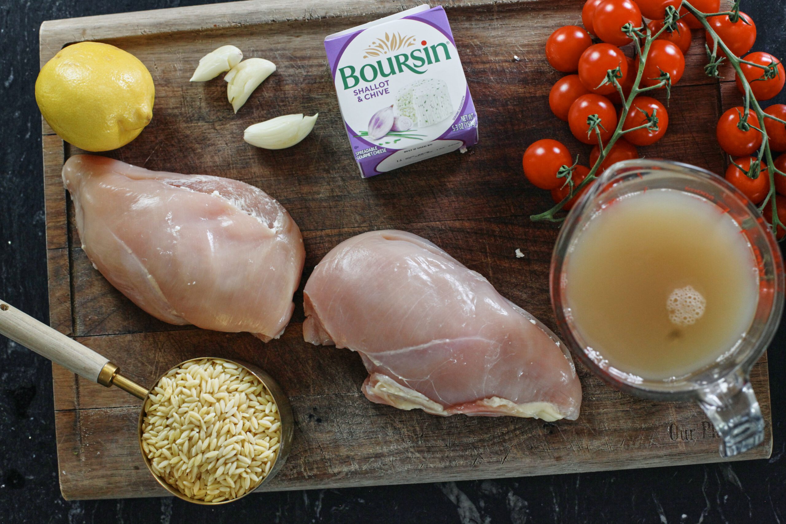 ingredients prepped and ready on a wooden cutting board: zested lemon, raw chicken breasts, orzo in a measuring cup, chicken broth in a glass measuring cup, cherry tomatoes, Boursin cheese, and garlic cloves