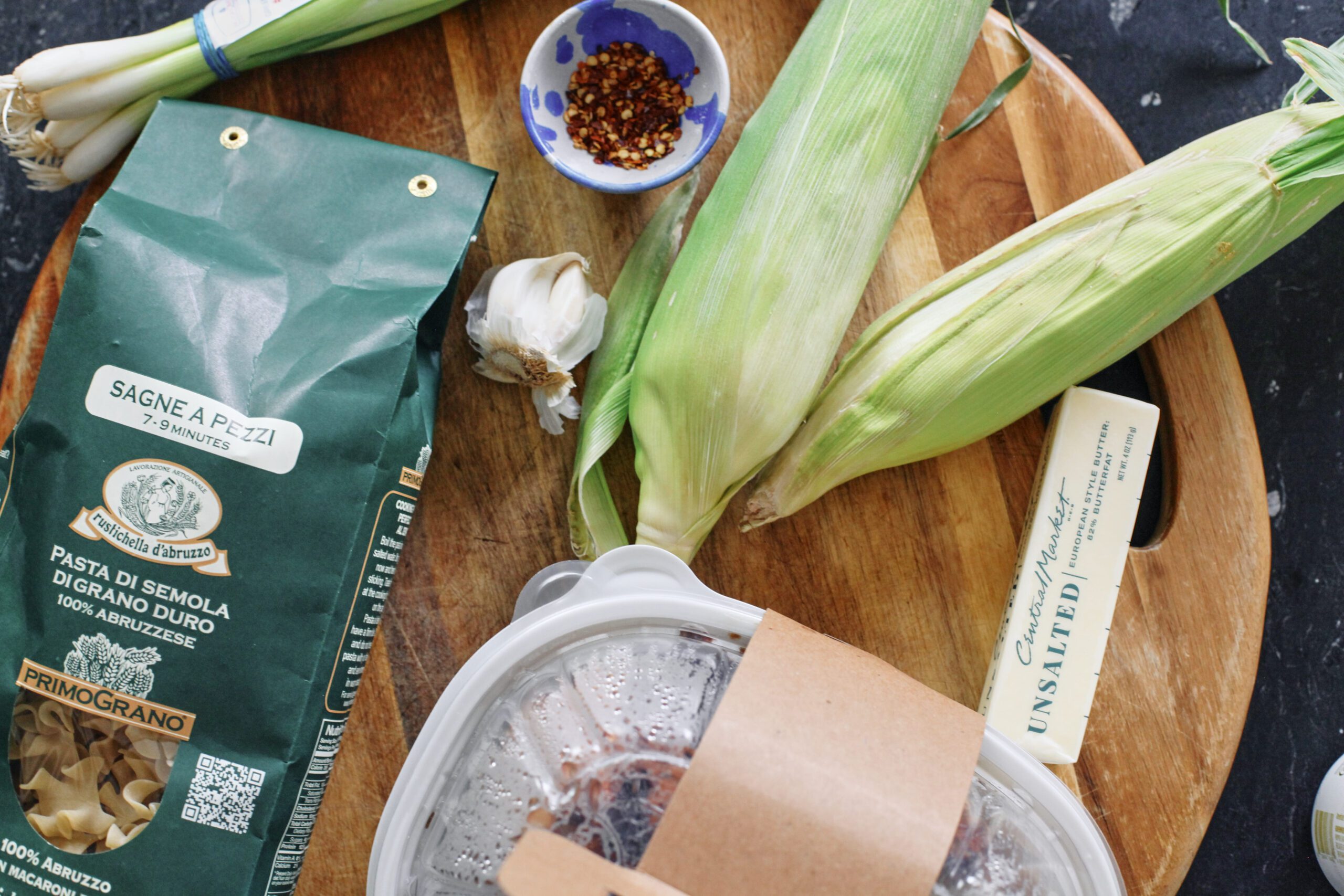 ingredients for the dish: bag of pasta, ears of corn, bunch of green onions, bulb of garlic, chili flakes in a bowl, rotisserie chicken in a plastic container, and a stick of butter