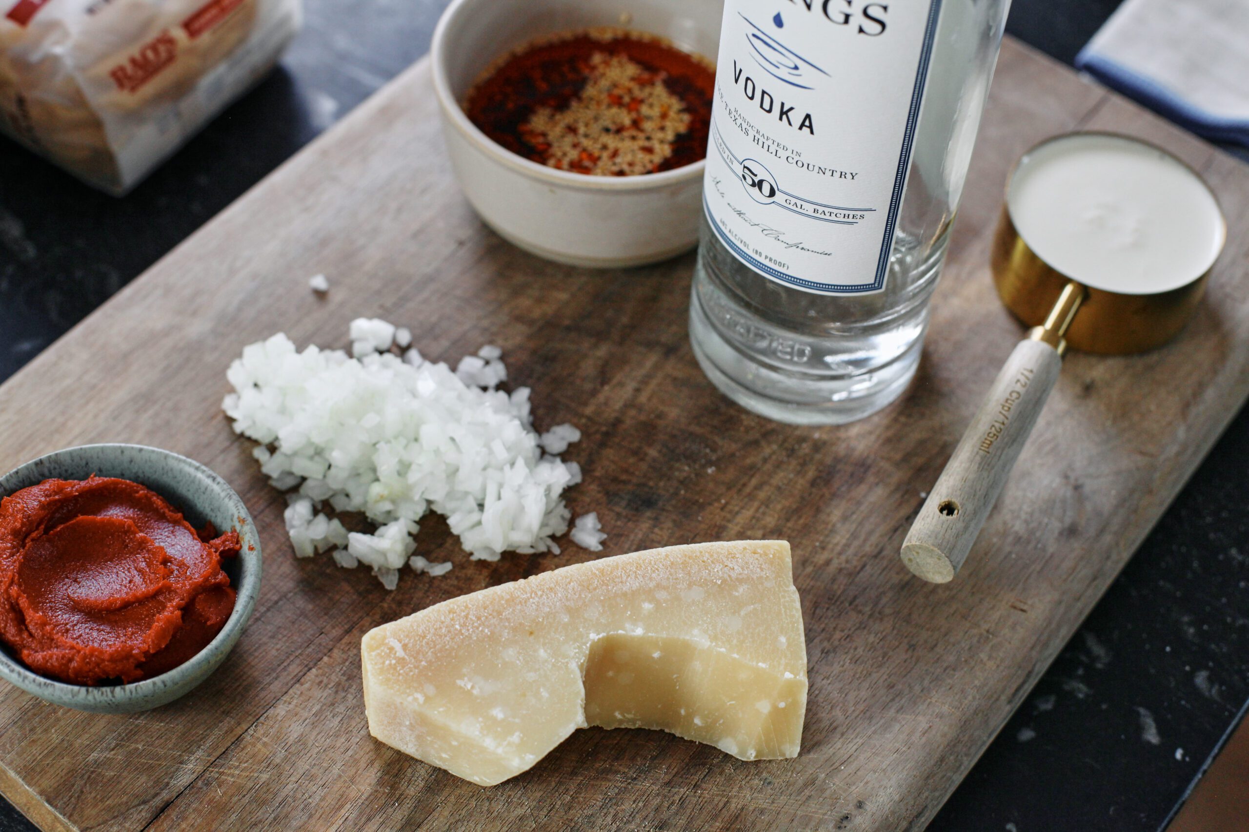 chili oil penne alla vodka 1 Ingredients on a cutting board prepped for Chili Oil Penne alla Vodka: a bag of pasta, tomato paste in a bowl, chopped onion, a chunk of parmesan cheese, a bowl of chili oil, a bottle of vodka, and heavy cream in a measuring cup.