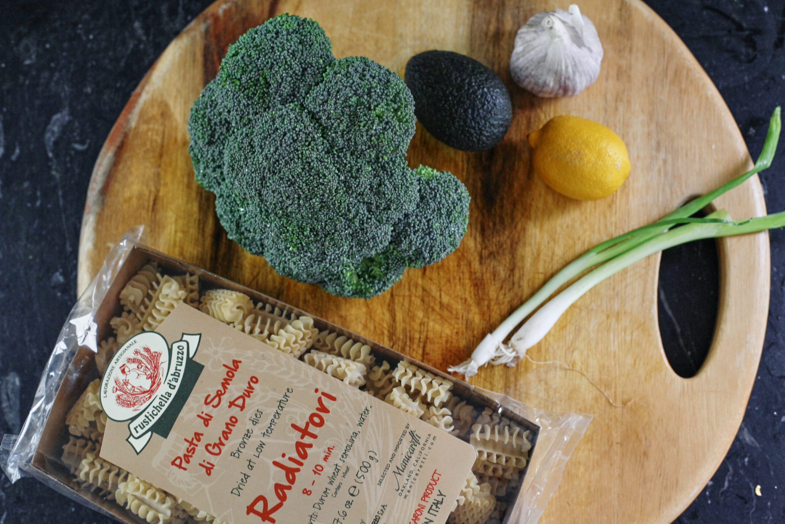 some of the ingredients for broccoli caesar pasta salad on a wood cutting board: pasta, broccoli, avocado, garlic, lemon, green onions