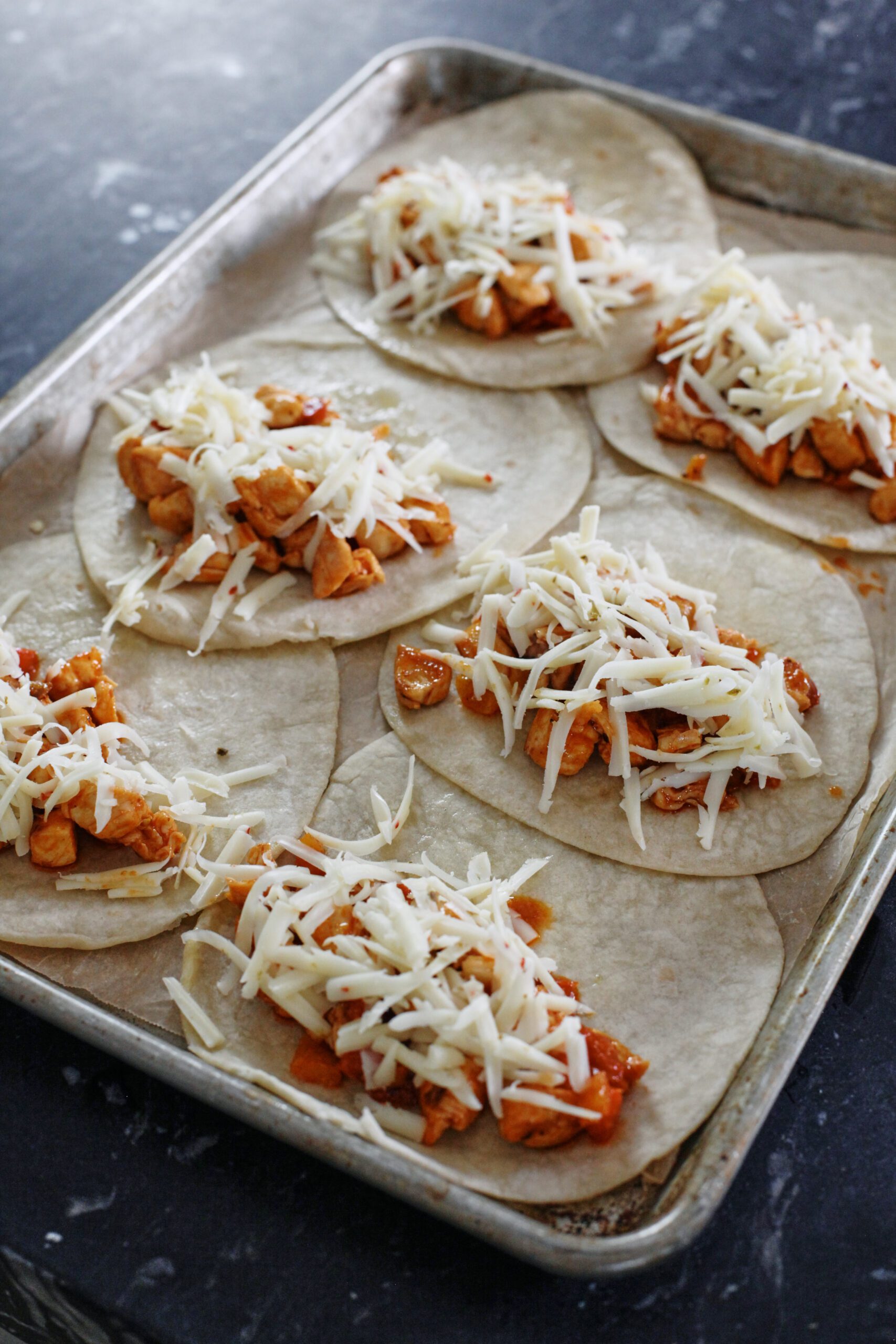tortillas on a sheet pan with the buffalo chicken mixture and shredded cheese on one half of each