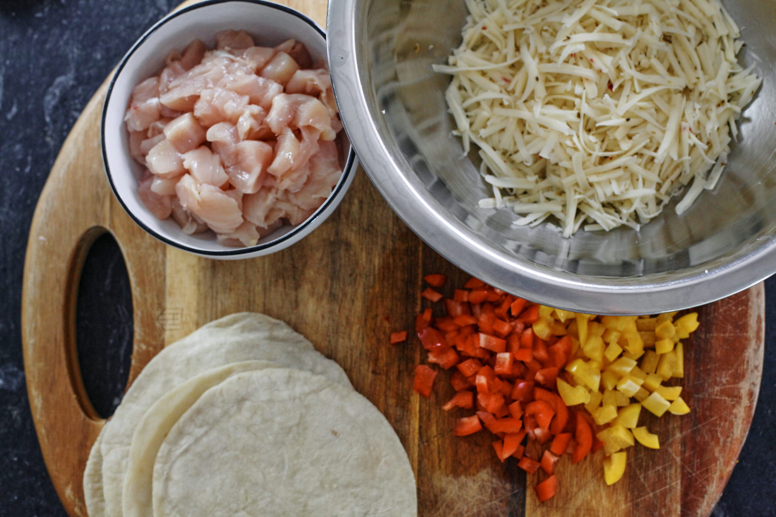 ingredients for buffalo chicken sheet pan tacos prepped on a wood cutting board: chopped raw chicken breasts in a bowl, tortillas, chopped yellow and red bell pepper, shredded cheese in a metal bowl