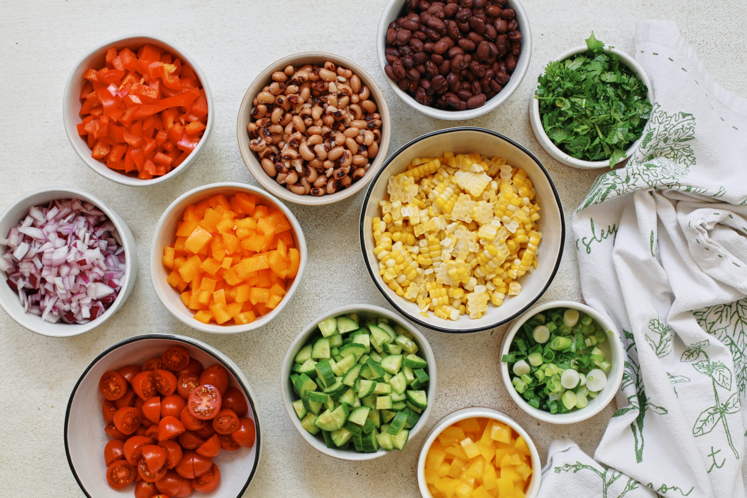 prepped ingredients for cowboy caviar pasta salad: bell peppers, cucumber, red onion, corn, cherry tomatoes, green onions, cilantro, black beans, black-eyed peas
