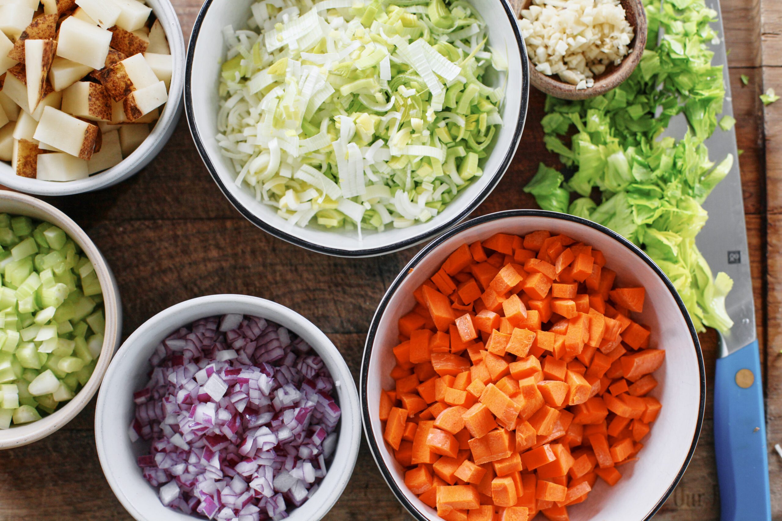 prepped ingredients in bowls on a wood cutting board: chopped carrots, red onion, celery, potato, leeks, garlic, and then celery leaves on the cutting board