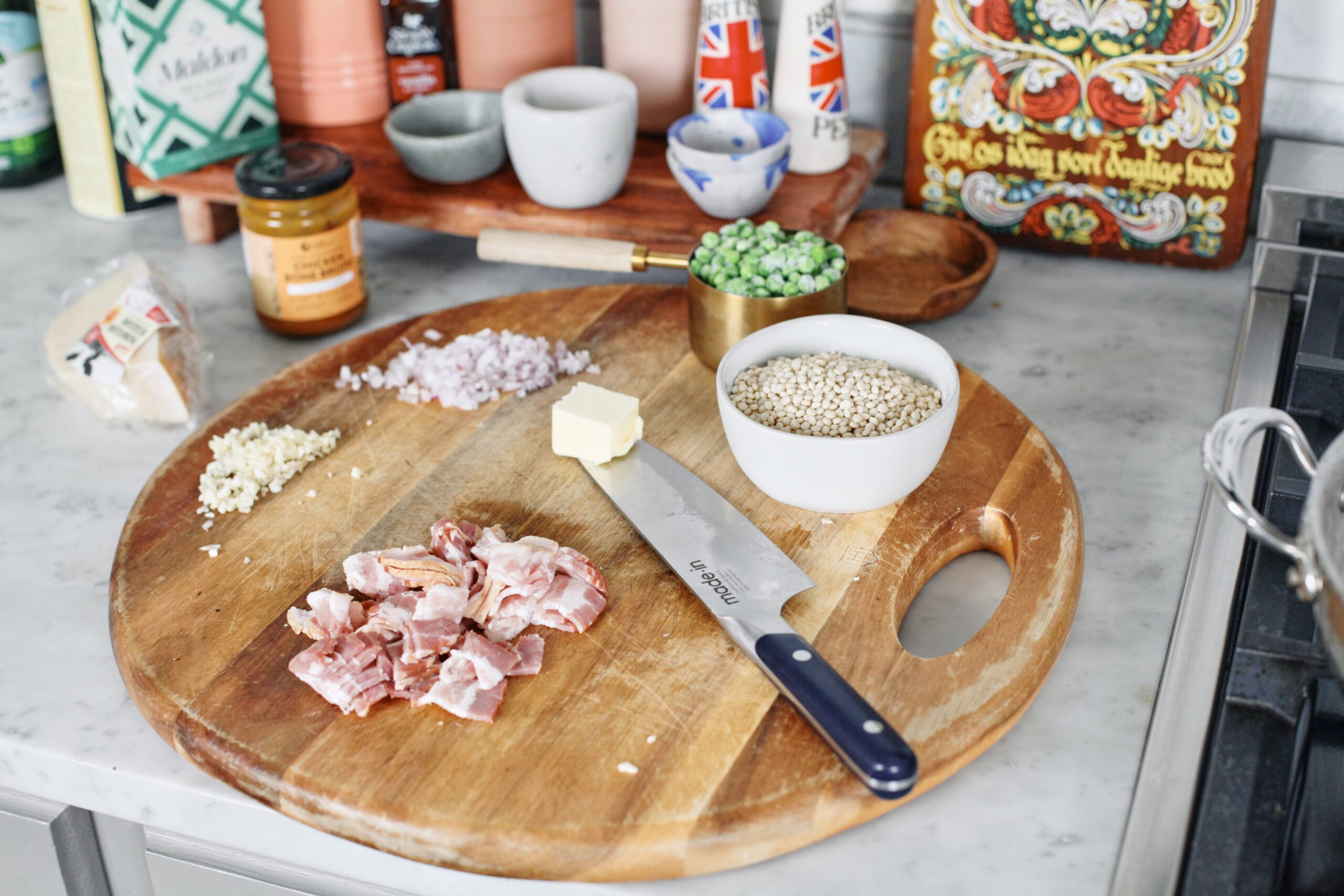 prepped ingredients on a round wooden currint board: minced shallot, minced garlic, chopped bacon, a cup of peas, a bowl of pearl couscous, a knob of butter on a knife