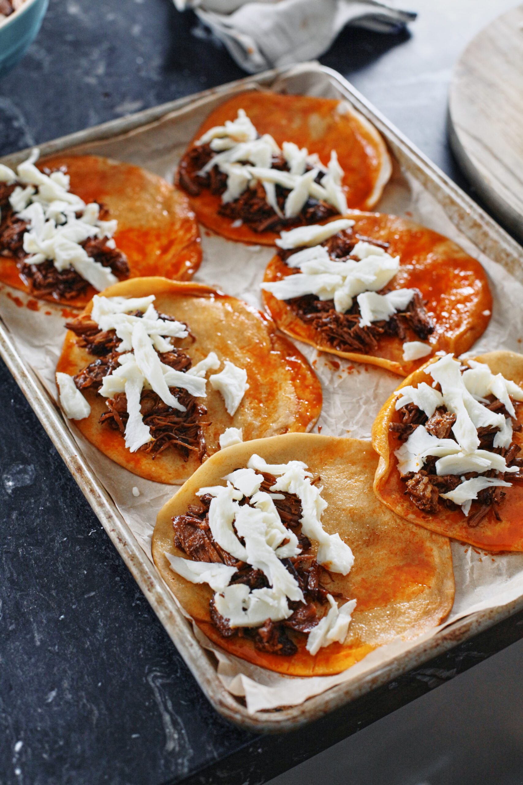 tortillas arranged on a parchment-lined sheet pan after being dipped in the birria consomme. one half of each tortilla has torn Oaxaca cheese and shredded beef