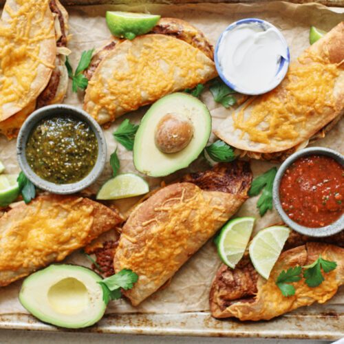 black bean sheet pan tacos arranged on a sheet pan with bowls of red and green salsa, lime wedges, a halved avocado, and cilantro
