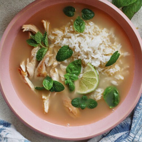 Overhead photo of a bowl of ginger chicken rice soup with shredded rotisserie chicken, fluffy white rice, and fresh mint and basil on top, garnished with a lime wedge