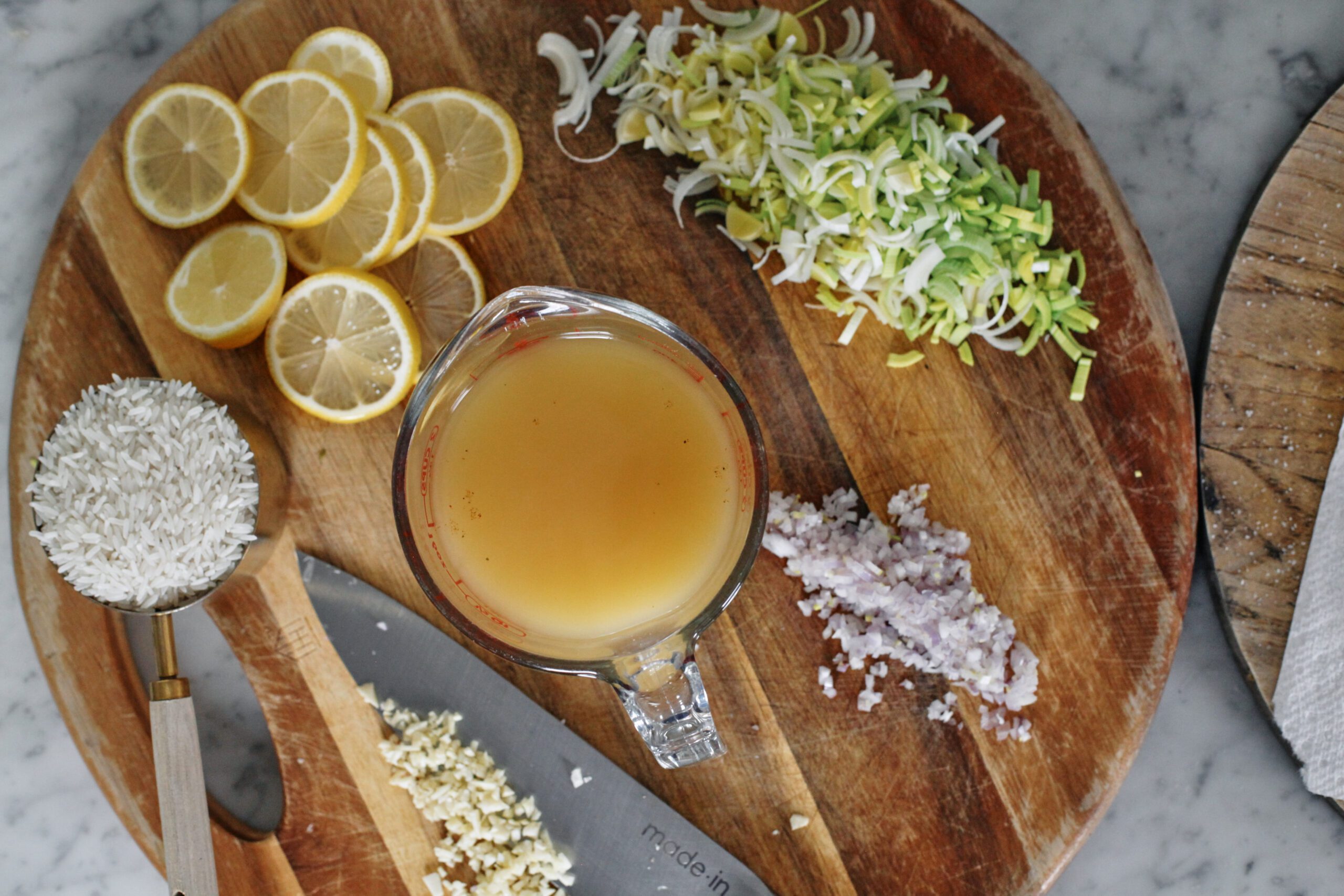 prepped ingredients on a round cutting board: a cup of white rice, lemon slices, minced garlic on a knife, a glass measuring container of broth, a chopped shallot, a minced shallot