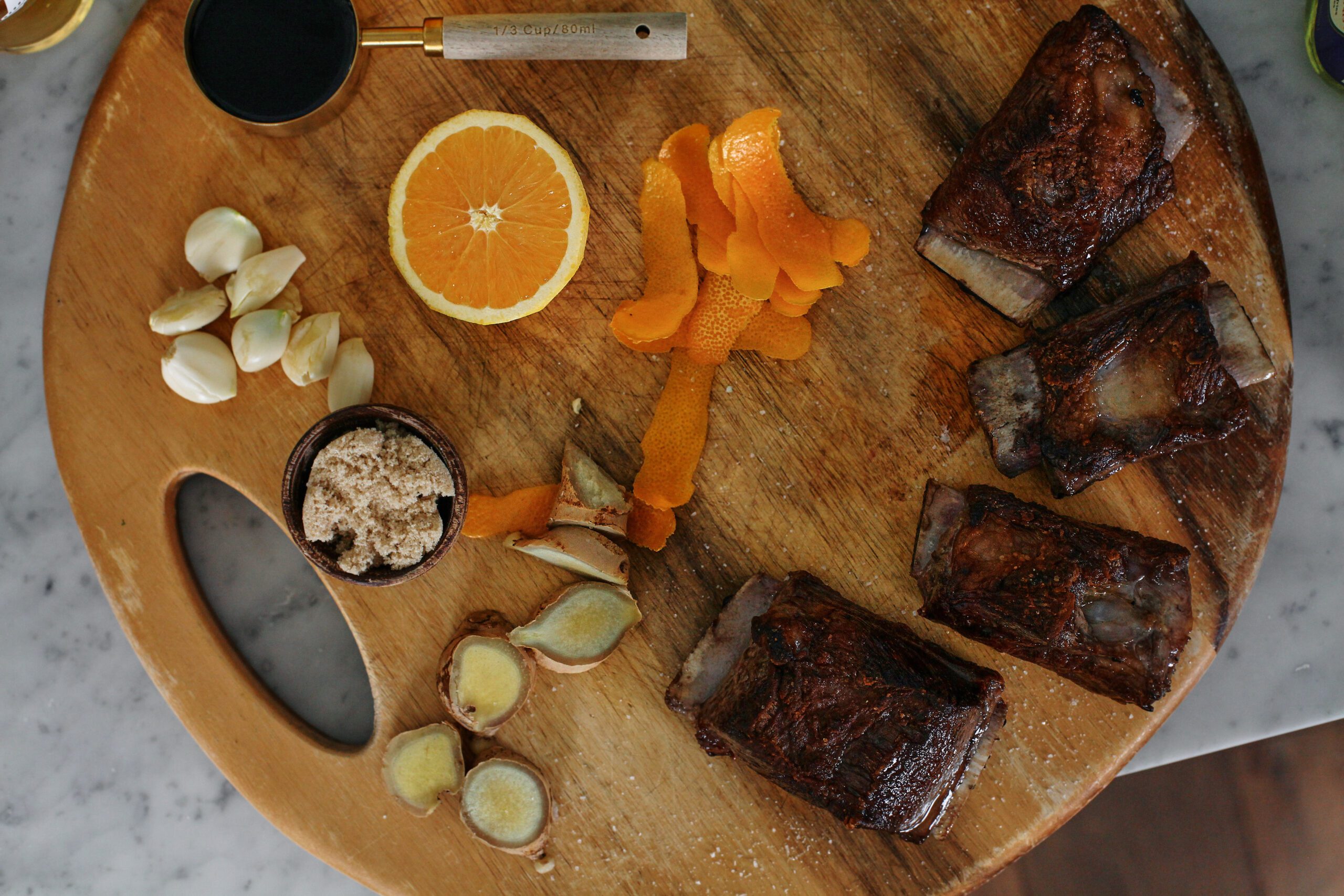 prepped ingredients: garlic cloves, orange peels, an orange, 1/2 cup of soy sauce, ginger sliced into coin-shaped pieces, seared short ribs