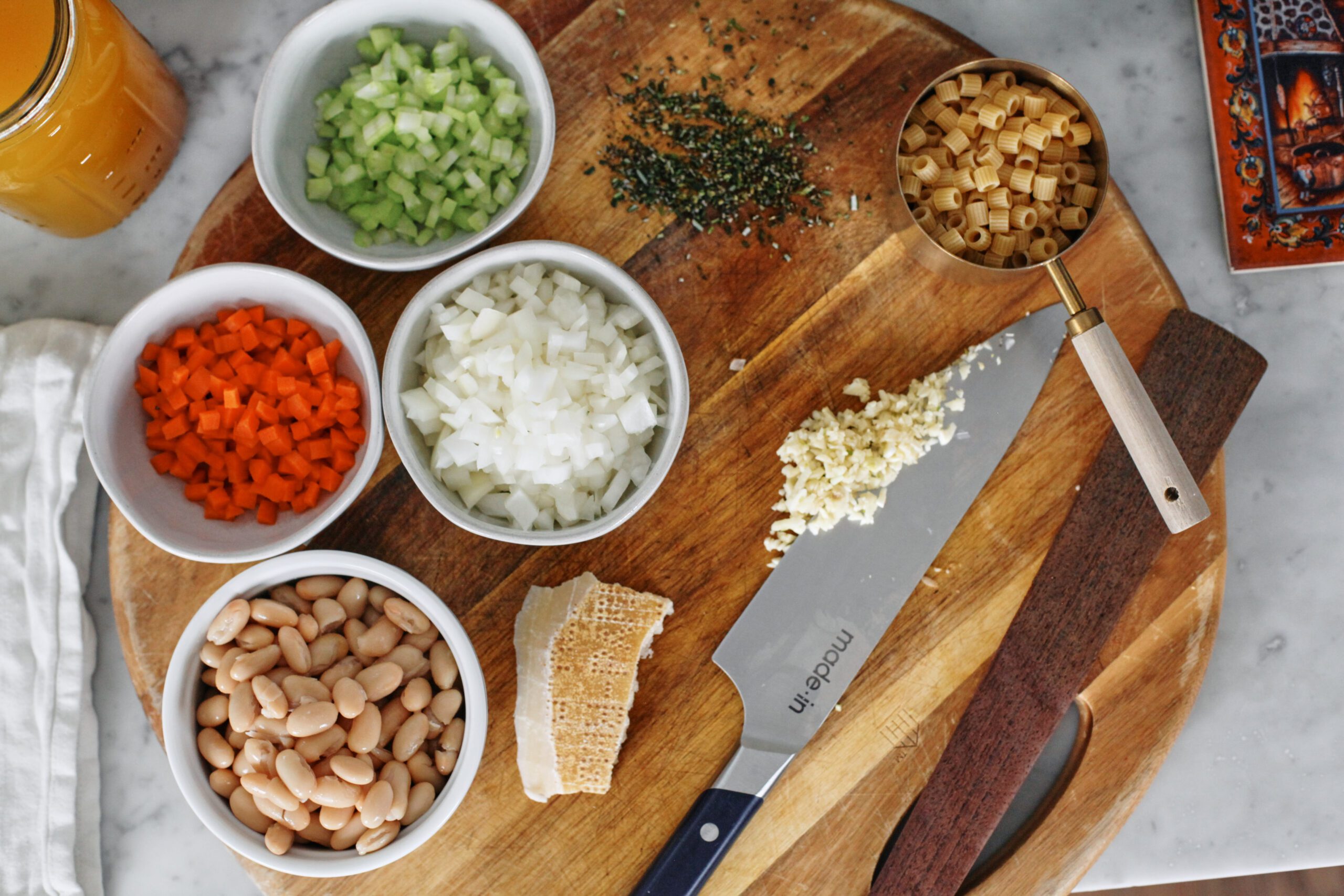 overhead photo of prepped ingredients on a round wooden cutting board: small white bowls of chopped celery, carrot, and onion, a small bowl of rinsed/drained white beans, a parmesan rind, minced garlic on the blade of a knife, minced fresh rosemary, and ditalini pasta in a measuring cup
