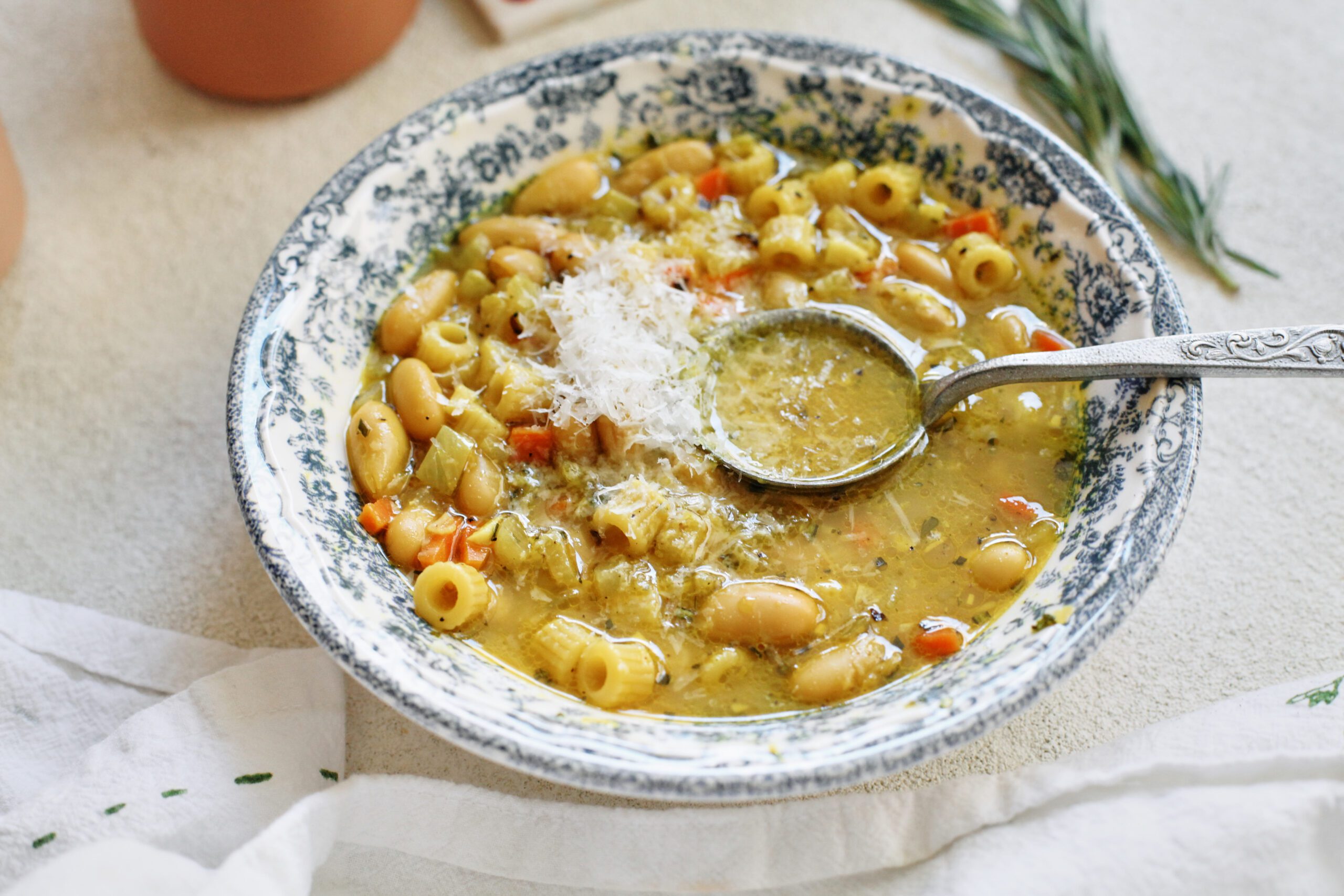 overhead photo of pasta e fagioli alla bianca in a blue/white floral patterned bowl. it's garnished with grated parmesan cheese and black pepper. an antique-looking silver spoon is in the bowl