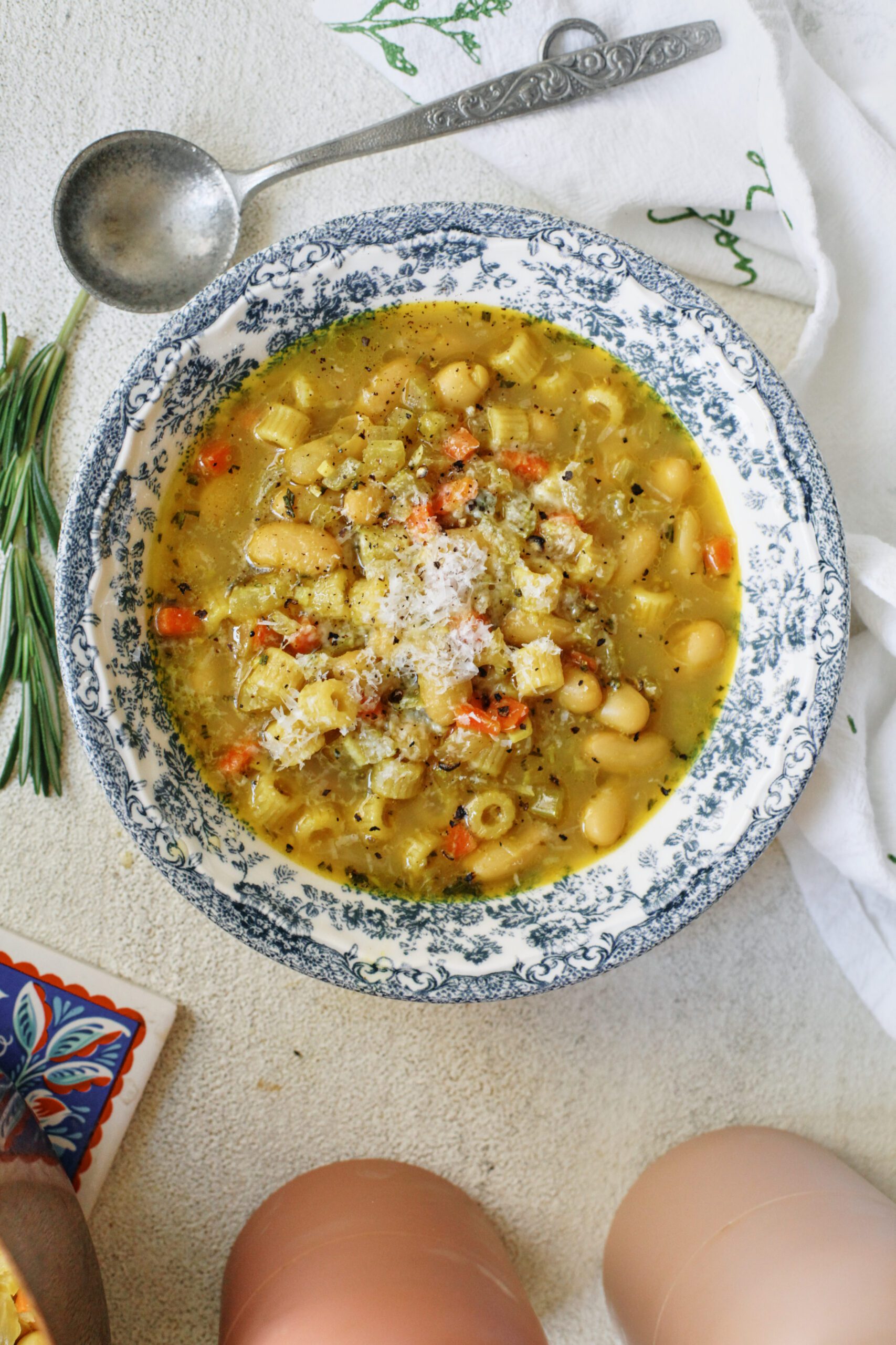 overhead photo of pasta e fagioli alla bianca in a blue/white floral patterned bowl. it's garnished with grated parmesan cheese and black pepper. an antique-looking silver spoon is next to the bowl