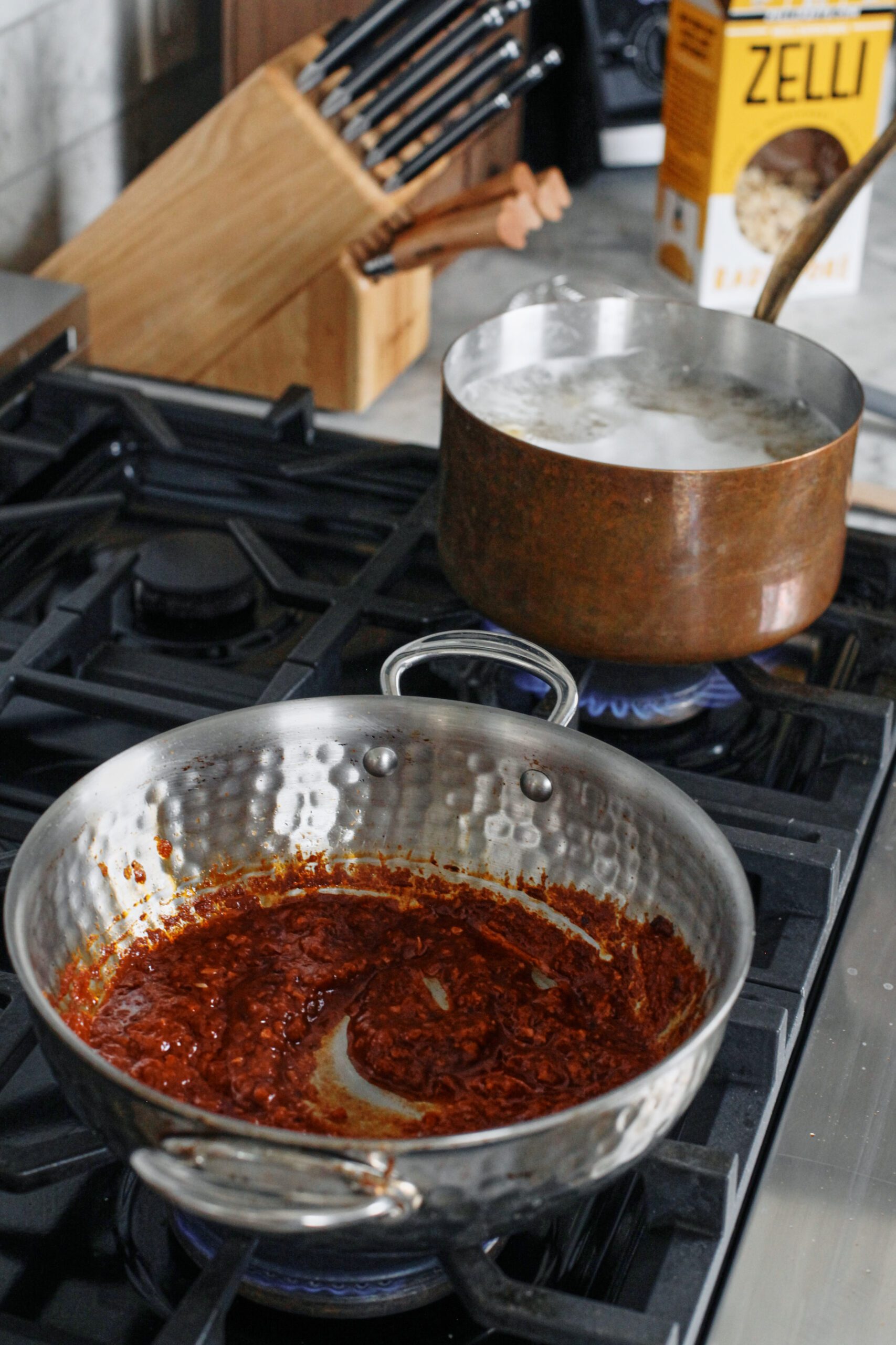 a hammered steel pan with the tomato paste mixture and a small bronze saucepan of boiling water on a gas range